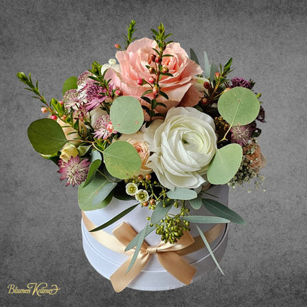 Elegante Blumenbox mit Rosen, Ranunkeln und Eukalyptus in Rosé- und Cremetönen in runder Geschenkbox mit Schleife